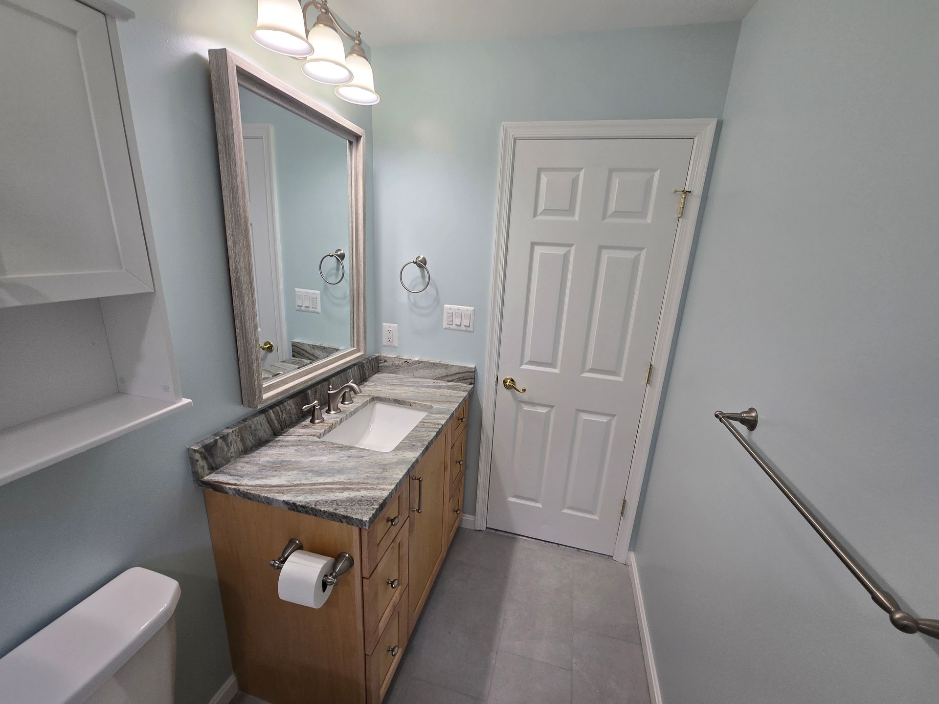 Bathroom with a vanity, toilet, and closed door; light blue walls and neutral floor.