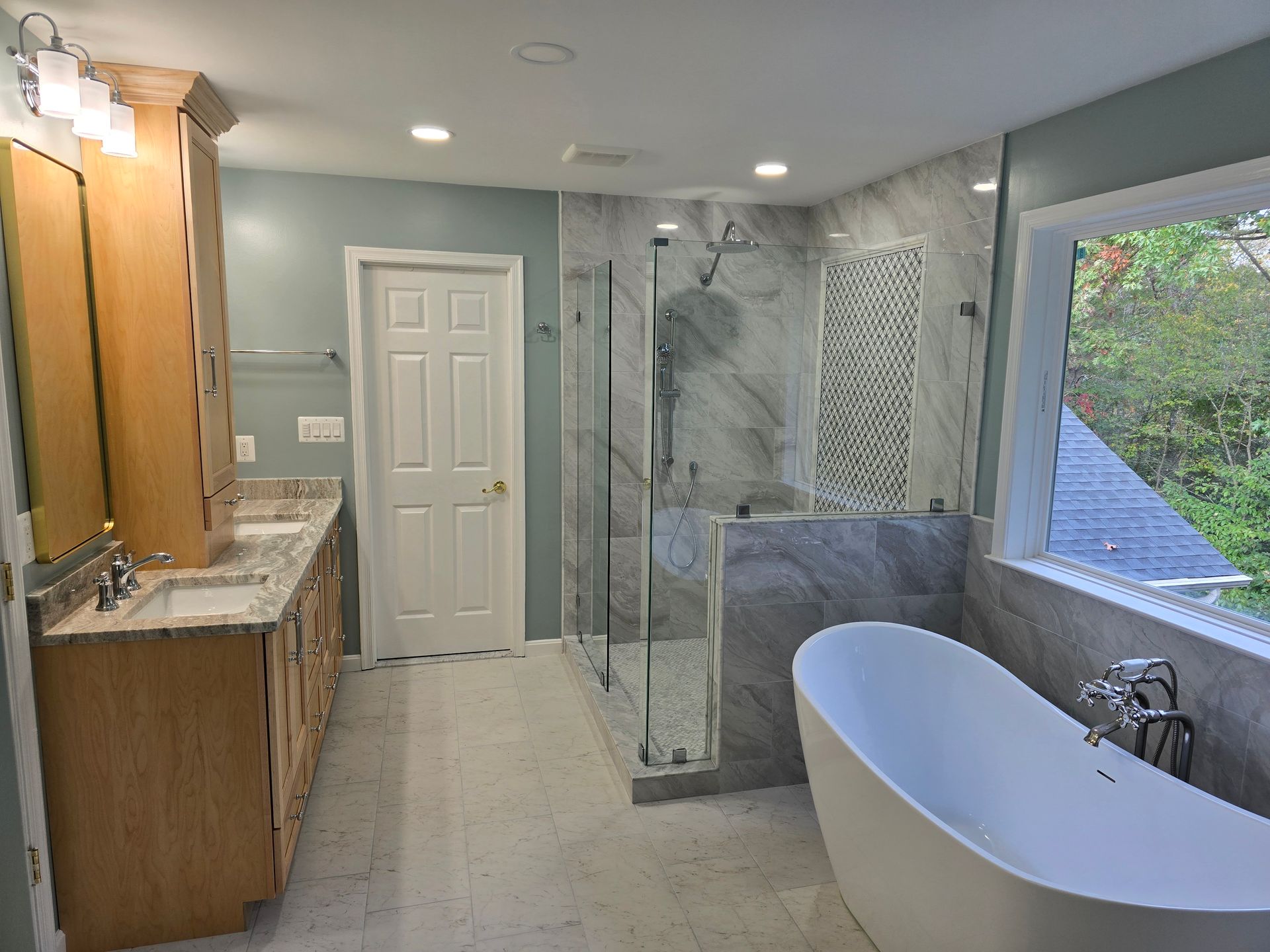 Spacious bathroom with light blue walls, marble tile shower, freestanding tub, double vanity, and a window.