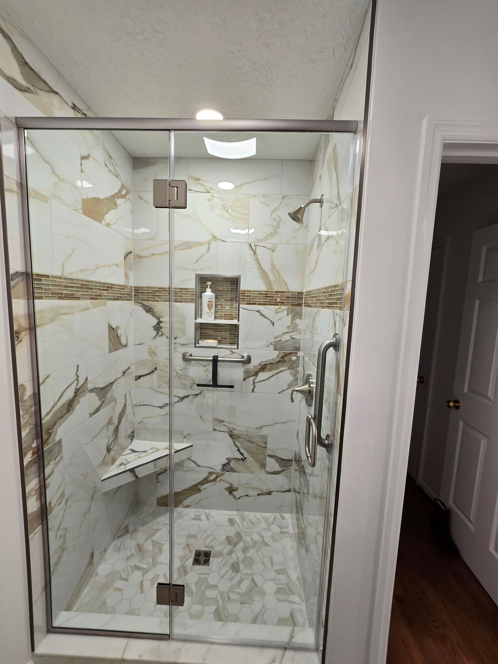 Shower with marble-like tile, glass doors, and a built-in bench. A doorway is visible on the right.