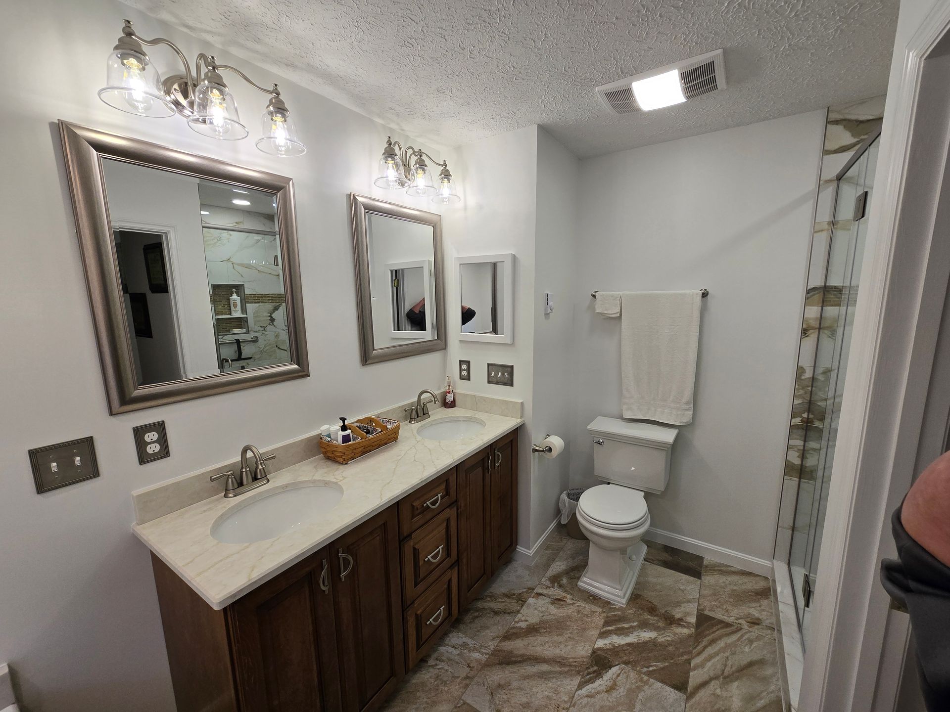 Bathroom with double sinks, mirrors, toilet, shower, and brown cabinets. Beige walls and patterned tile.
