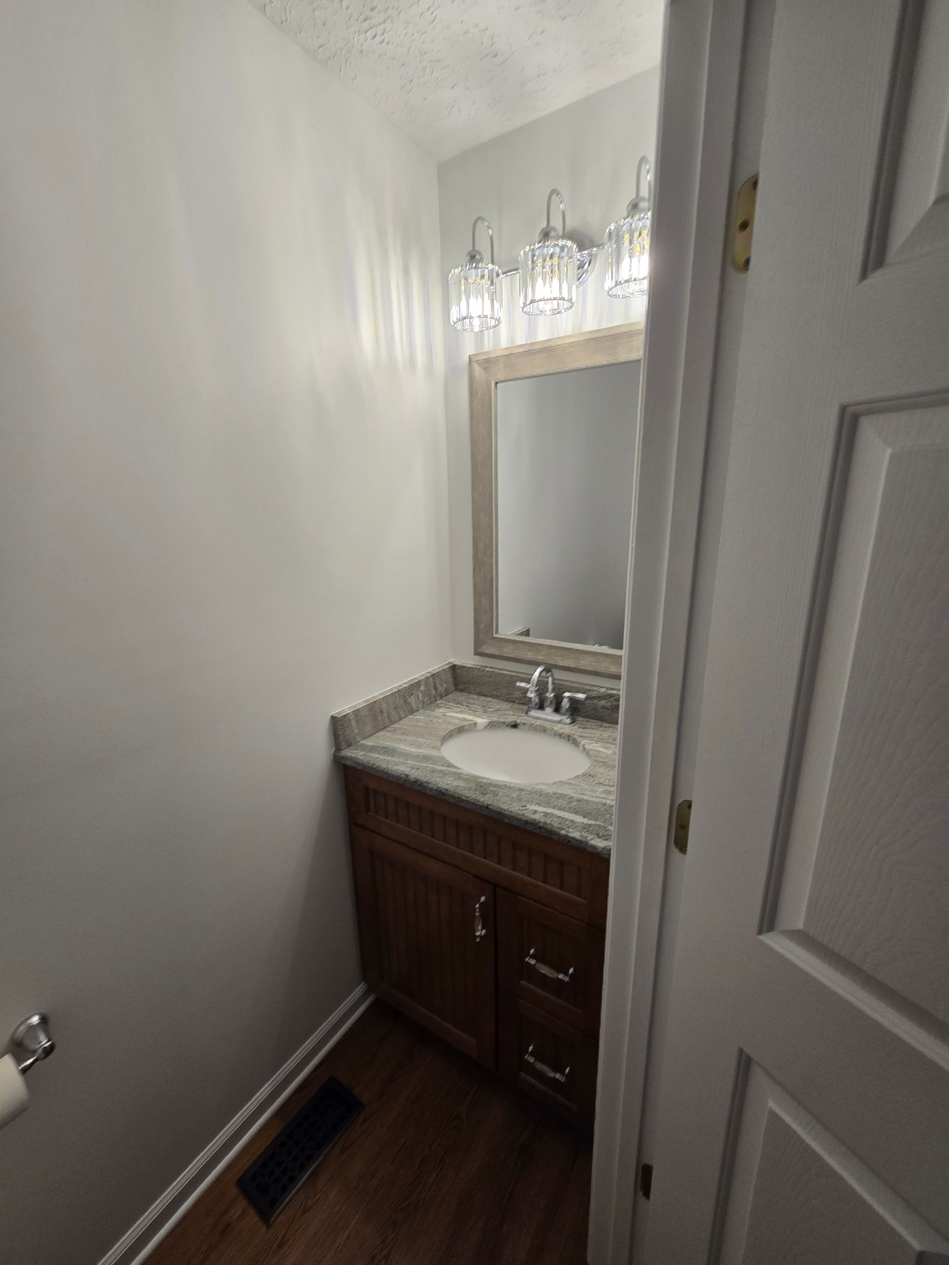 Small bathroom with vanity, mirror, and overhead lights; door frame visible on right.