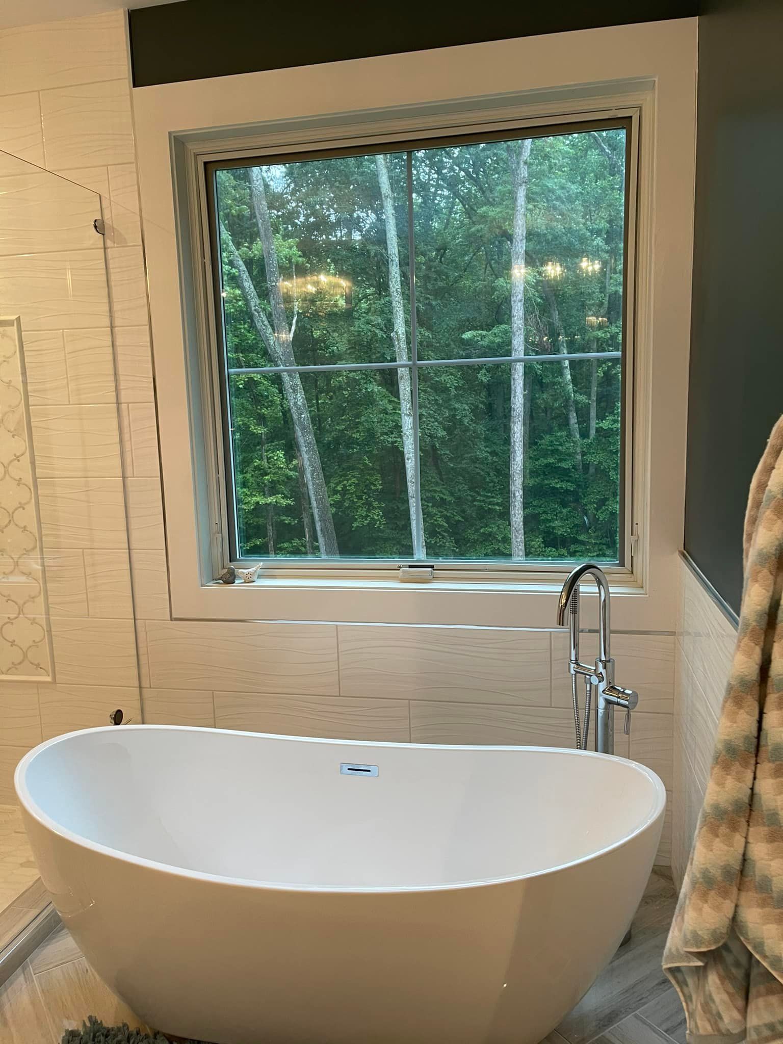White freestanding tub in a modern bathroom with a window overlooking trees.