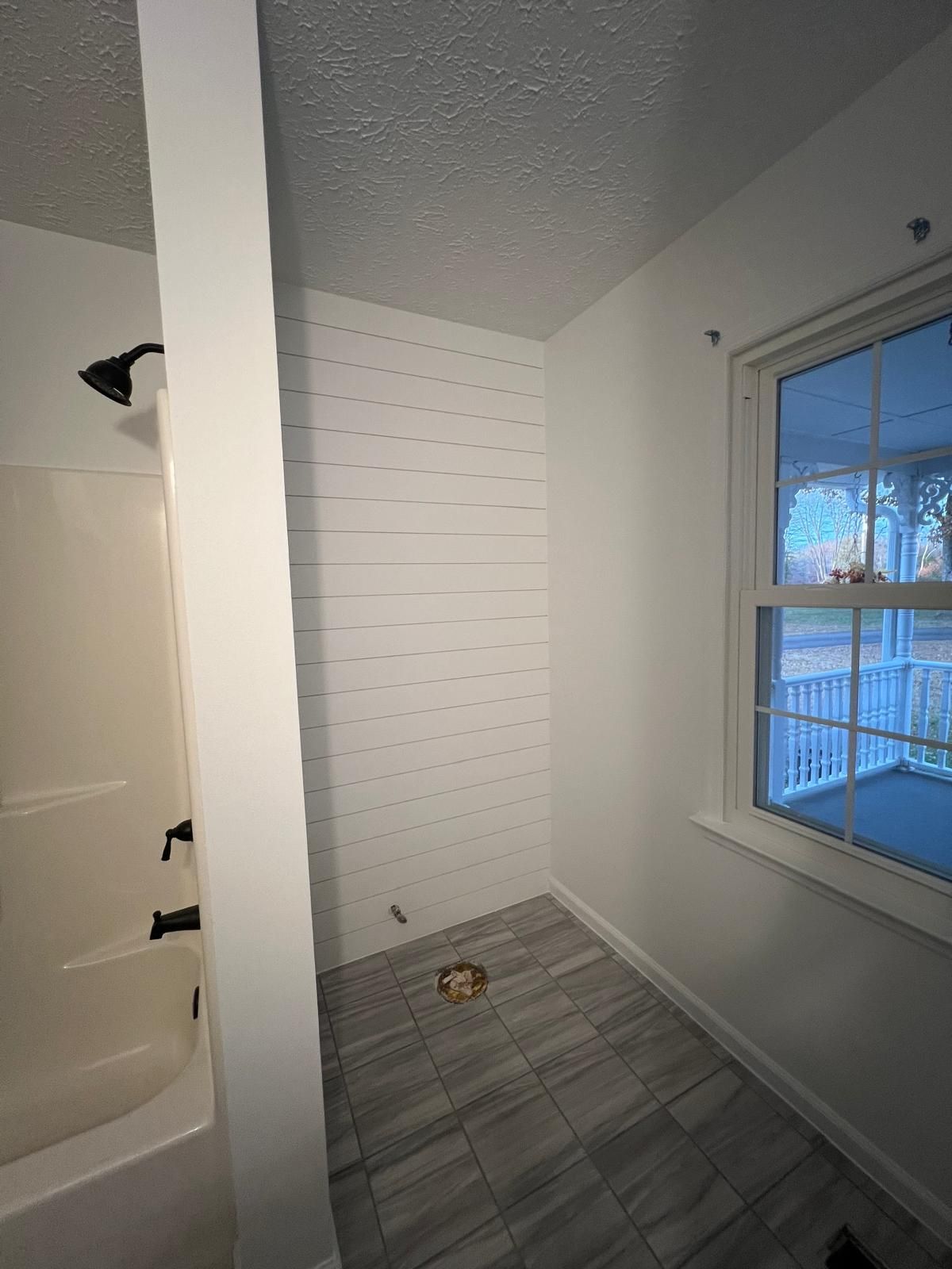 Bathroom with white shiplap wall, shower, window, and gray tiled floor.