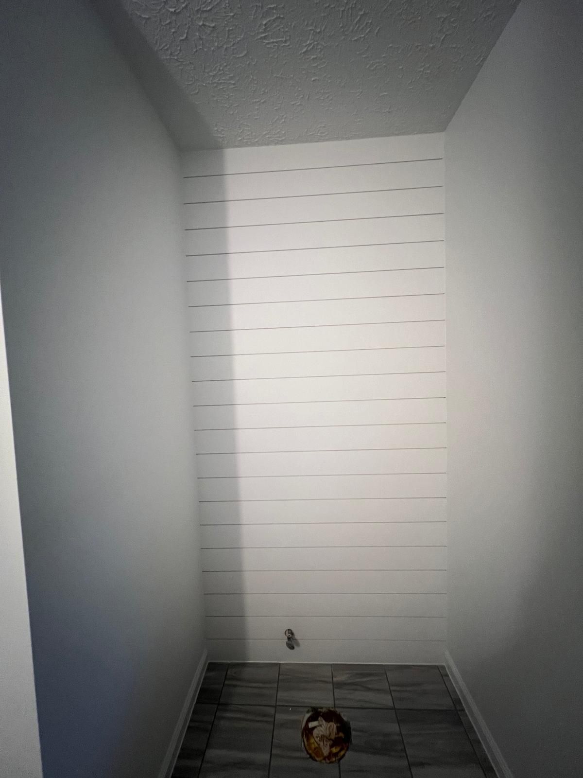 Small white-walled room with horizontal white plank accent wall, grey tiled floor, and a plumbing fixture.