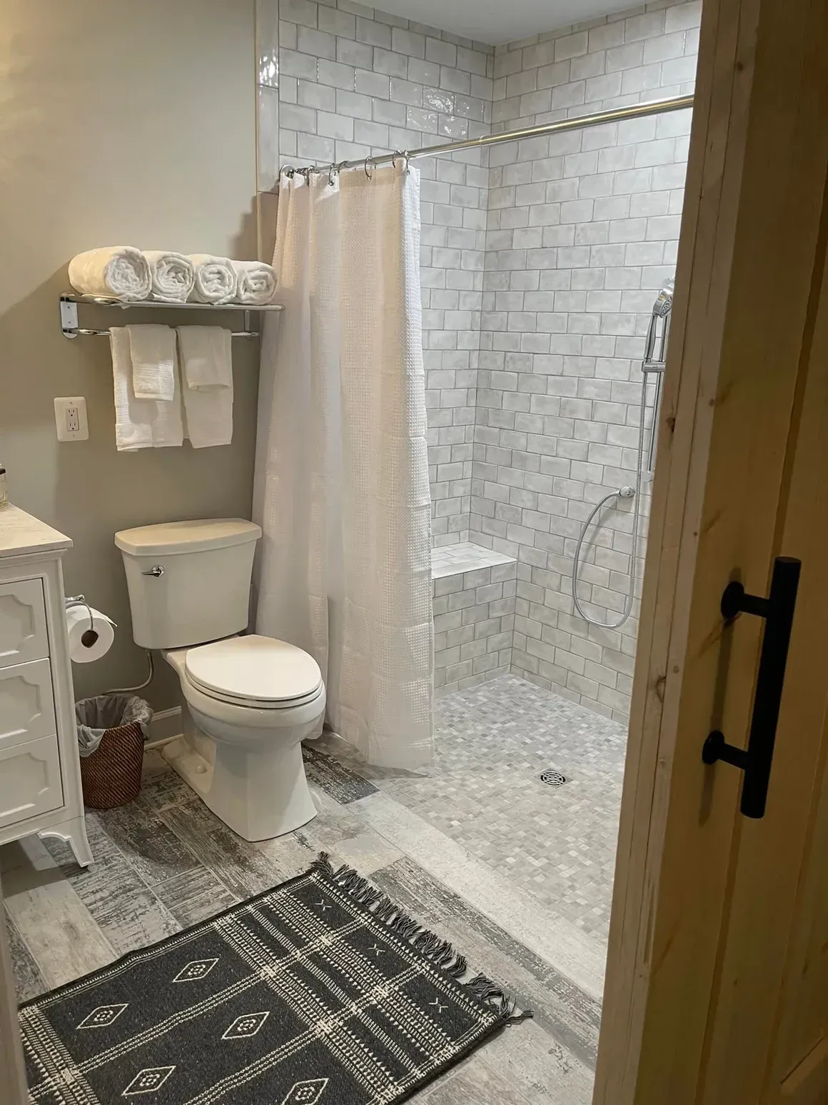 Bathroom with white tile shower, toilet, towels, and rug. Gray and white color scheme.
