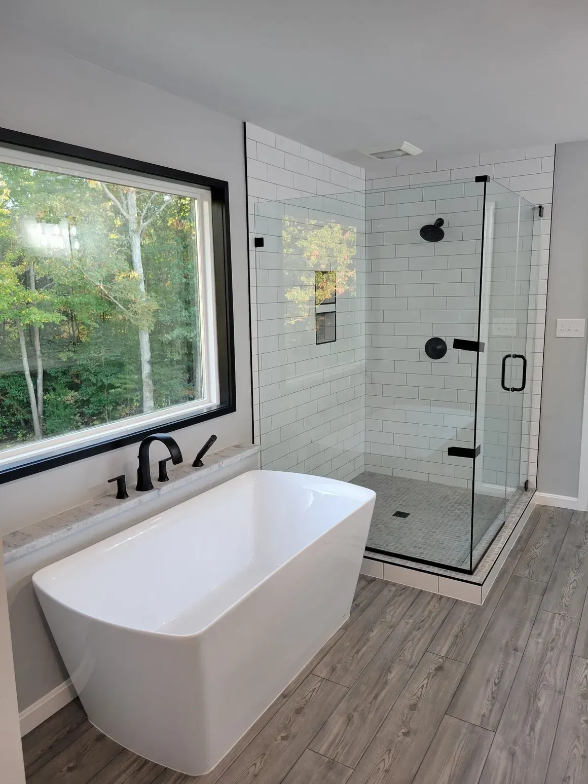 Modern bathroom with white tub, glass shower, and gray walls, with a window overlooking trees.