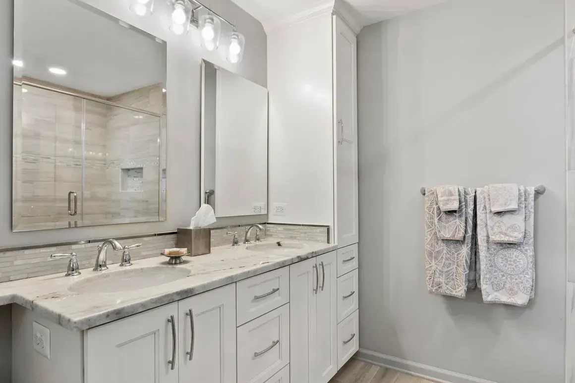 White bathroom with double vanity, large mirror, and shower; towels hang on wall.