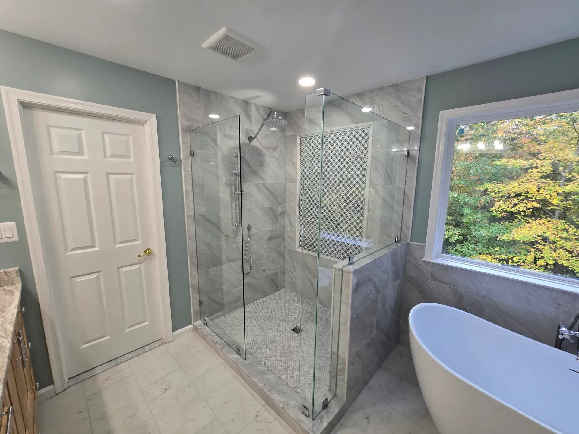 Bathroom with a glass-enclosed shower, freestanding tub, and a window overlooking trees; gray and white tile.