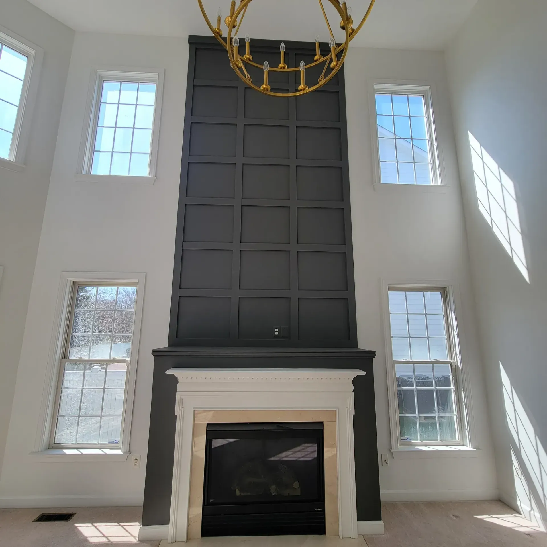 Gray fireplace and accent wall with four windows on white walls, gold chandelier.