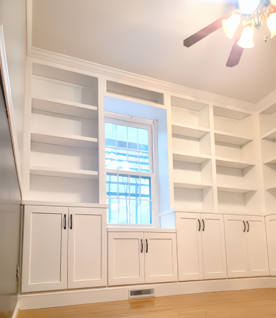 Built-in white bookshelves surround a window. Lower cabinets with black handles sit below the shelves.