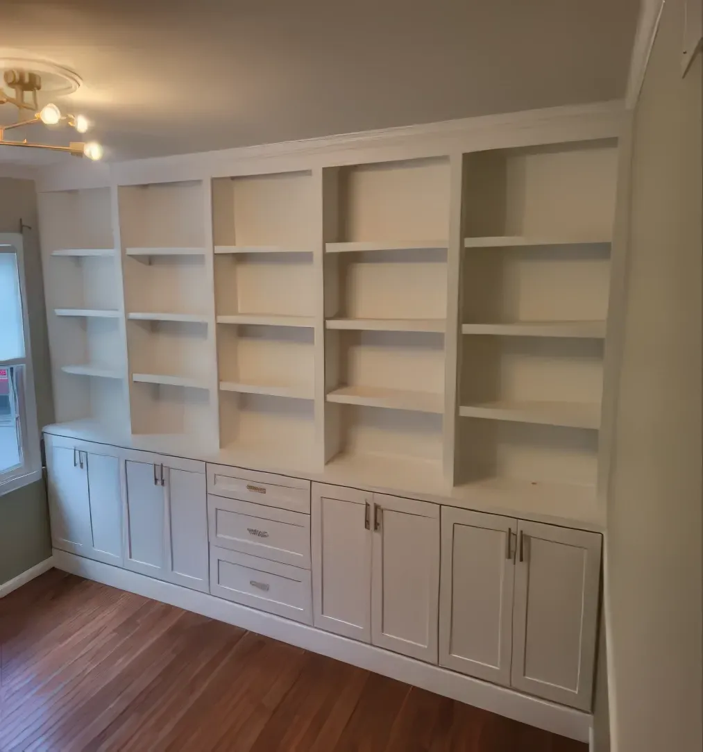 Built-in white bookshelves with cabinets, drawers, and open shelves, against a white wall in a room with wood flooring.