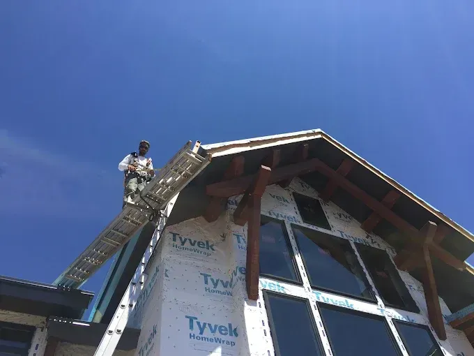 Man on a ladder working on a house roof under a blue sky. House has Tyvek wrap.