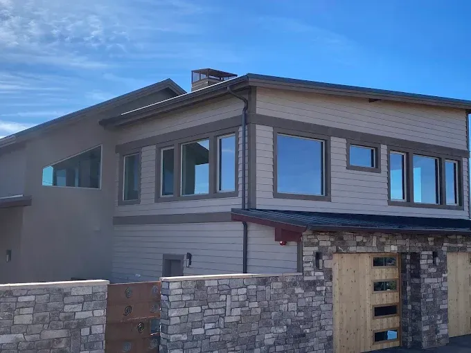 Two-story house with beige siding, brown trim, and large windows. Stone wall and gate in the foreground. Blue sky.