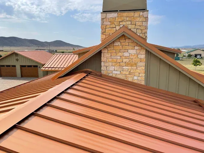 Copper-colored metal roof on a house with a stone chimney against a blue sky.