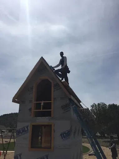 A person works on the roof of a small building. They're wearing a tool belt, under a bright sky.