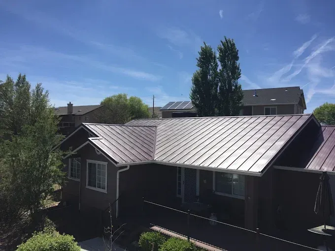 Brown house with metal roof under a bright blue sky, trees in the background.