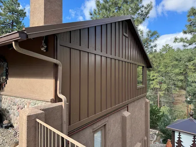 Brown siding on a house with a gutter and a small window, set against trees and a blue sky.