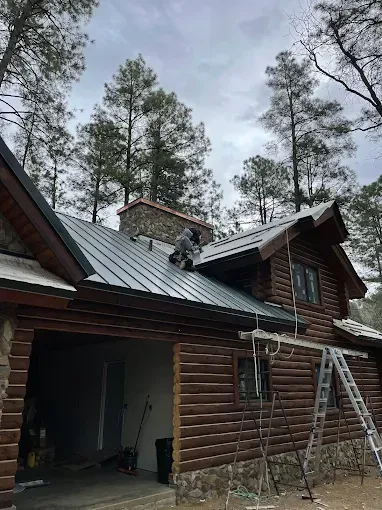 Person on a metal roof, working on a chimney of a log cabin with a garage. Cloudy sky, trees.