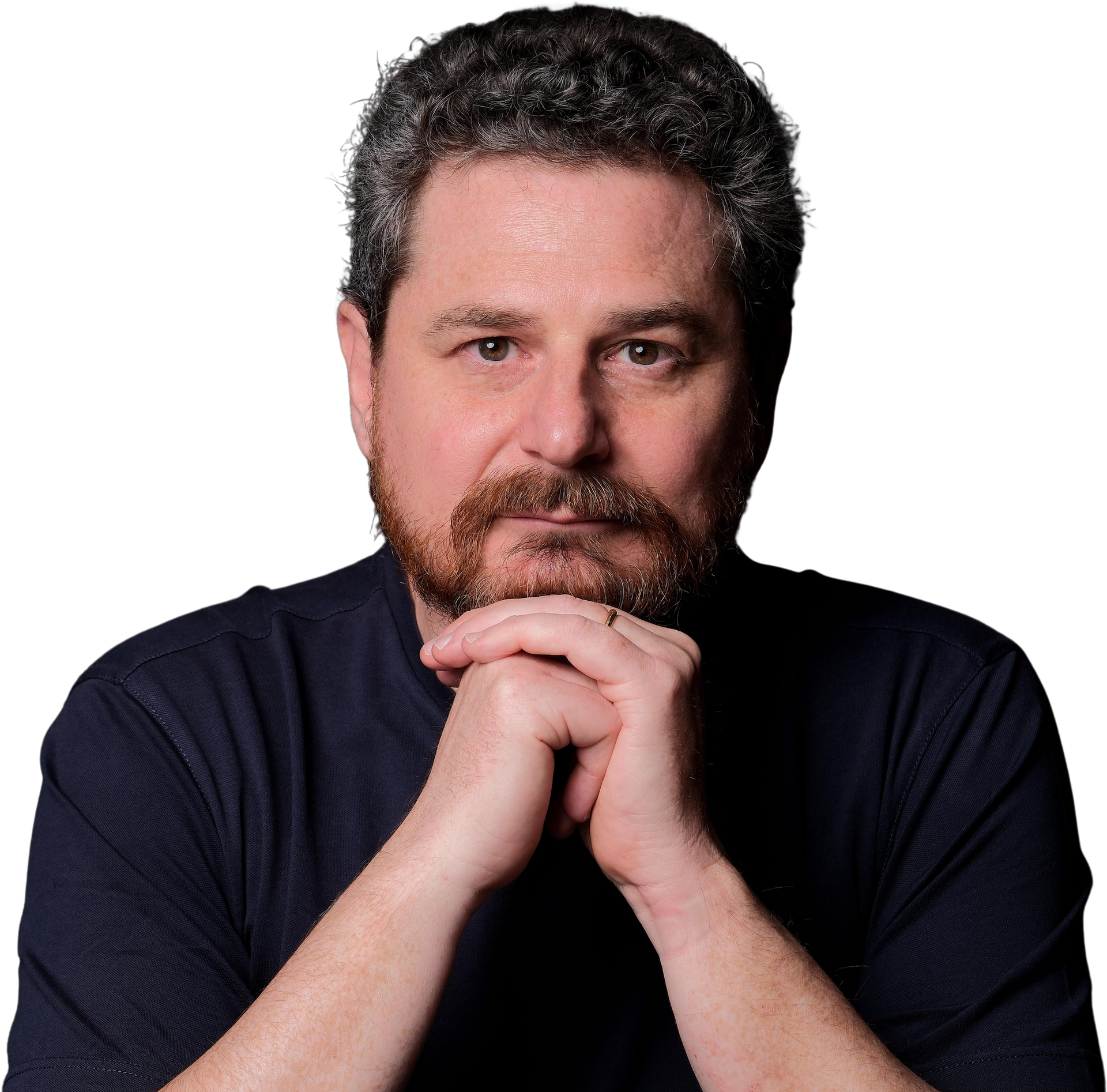 A person with short curly hair and a beard, wearing a dark shirt, resting their chin on clasped hands against a white background.