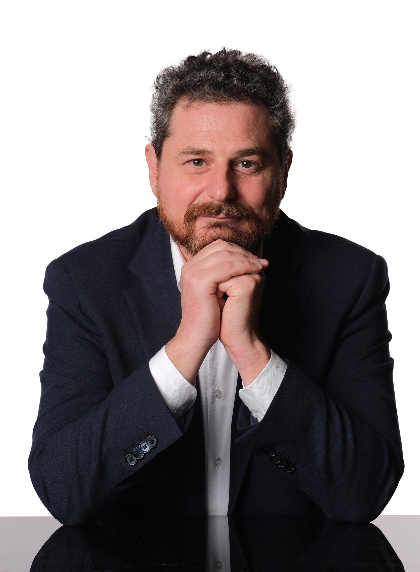 Man with beard in a suit, hands clasped under his chin, looking toward the viewer with a thoughtful expression.