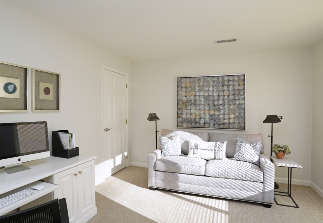 Home office with a white desk and sofa, a door, framed artwork and two lamps.