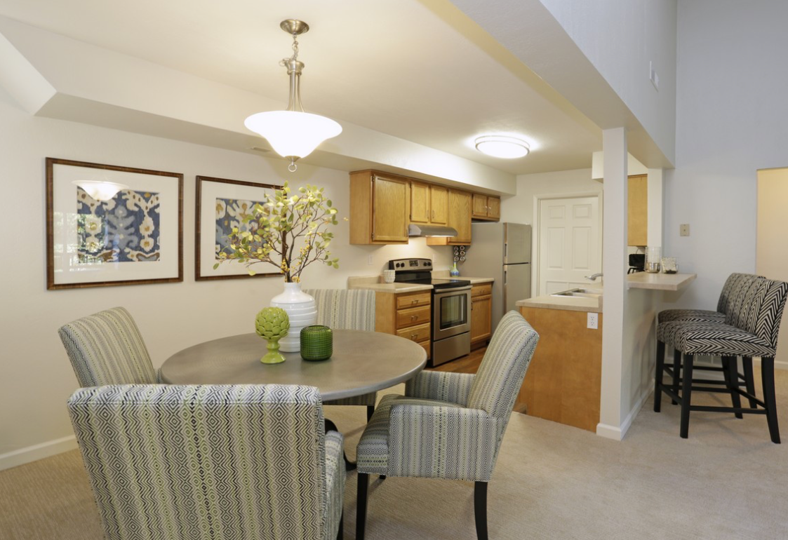 Dining area with table, chairs, and kitchen in the background. Beige and light wood tones.