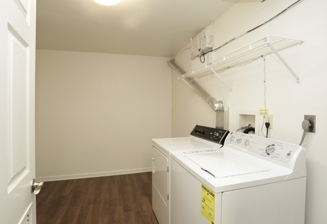 Laundry room with washer, dryer, drying rack, and door; hardwood floor and white walls.