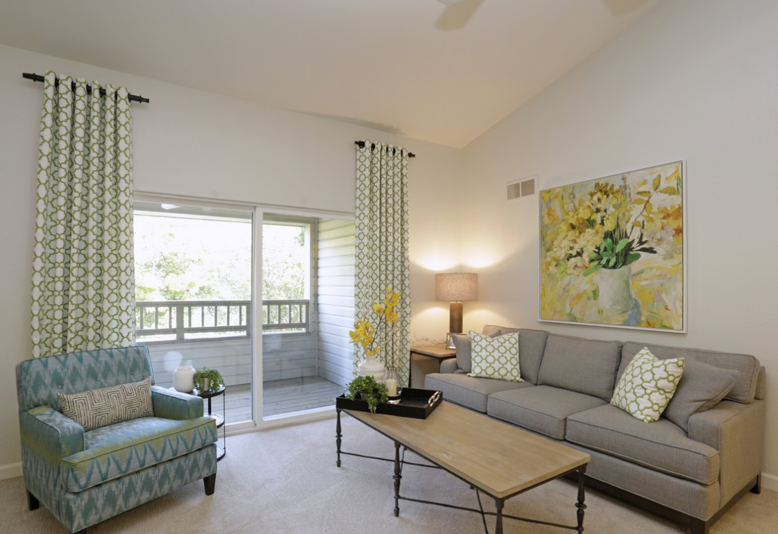 Living room with grey couch, teal armchair, yellow painting, and patterned curtains.