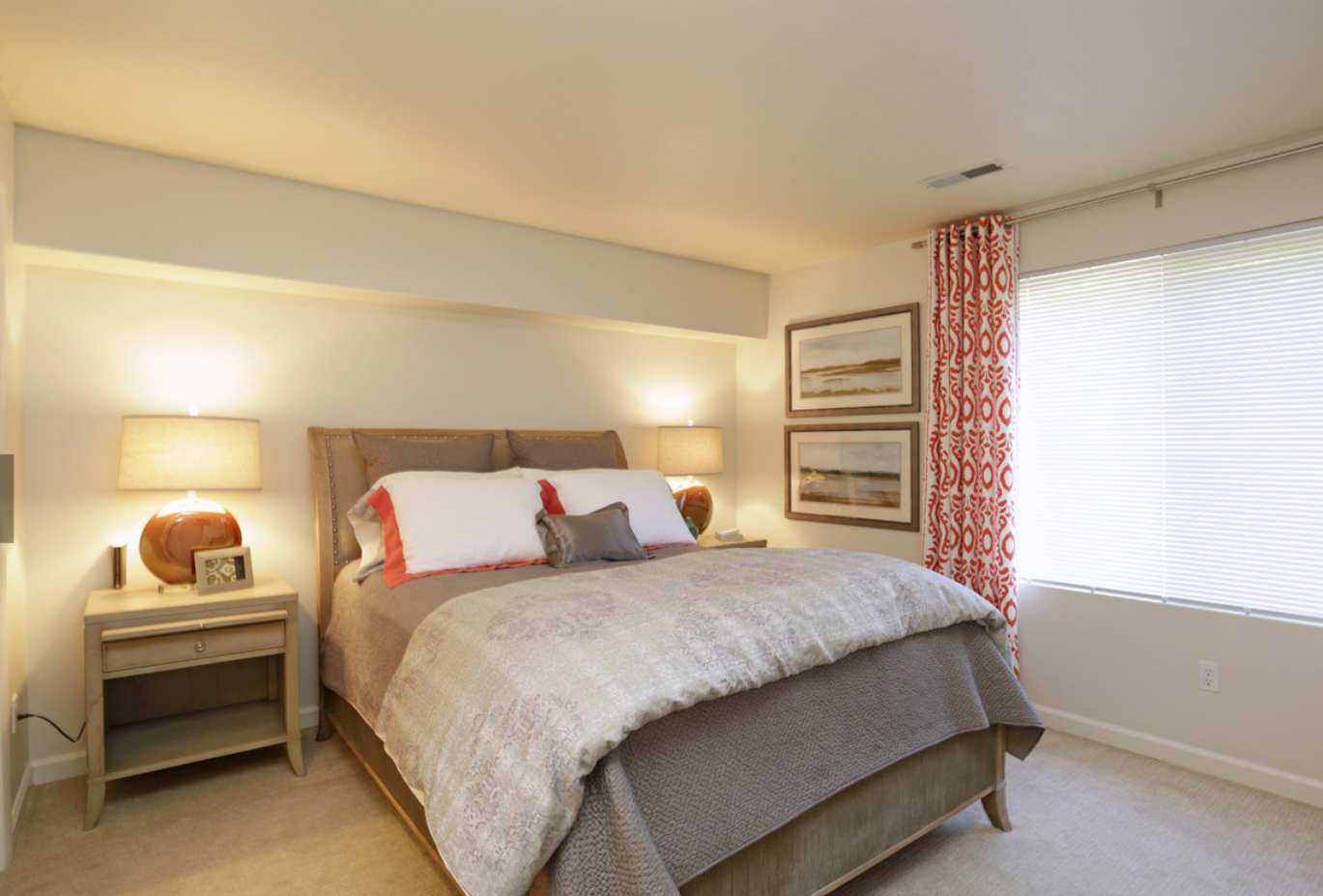 Bedroom with bed, nightstands, lamps, patterned bedding, and a window with blinds and patterned curtains.