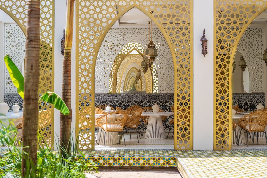 Ornate gold-trimmed archway with woven chairs and tables. Decorative screens and lanterns in a sunlit outdoor space.