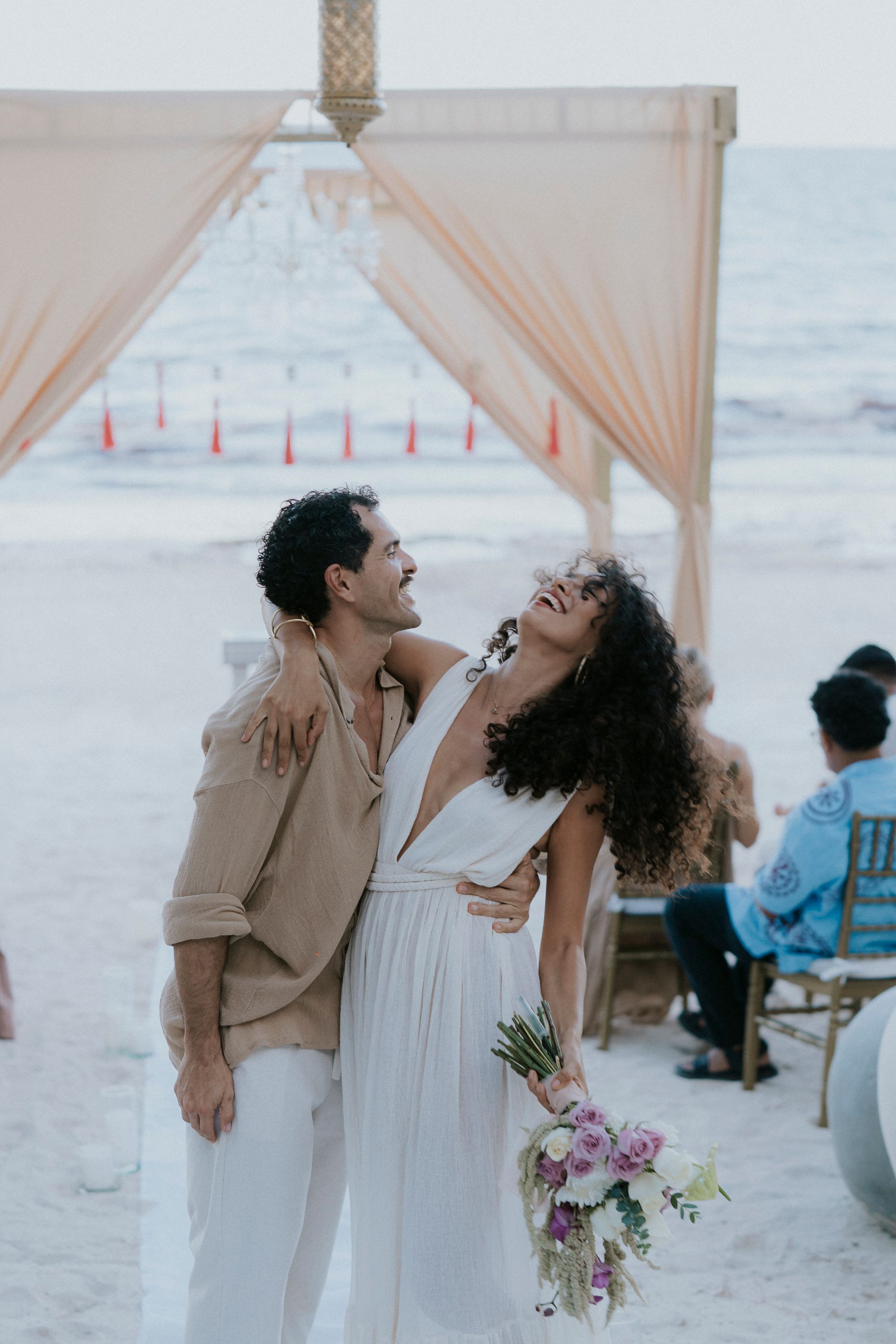 Couple in white outfits hold hands under an archway, beach in background.