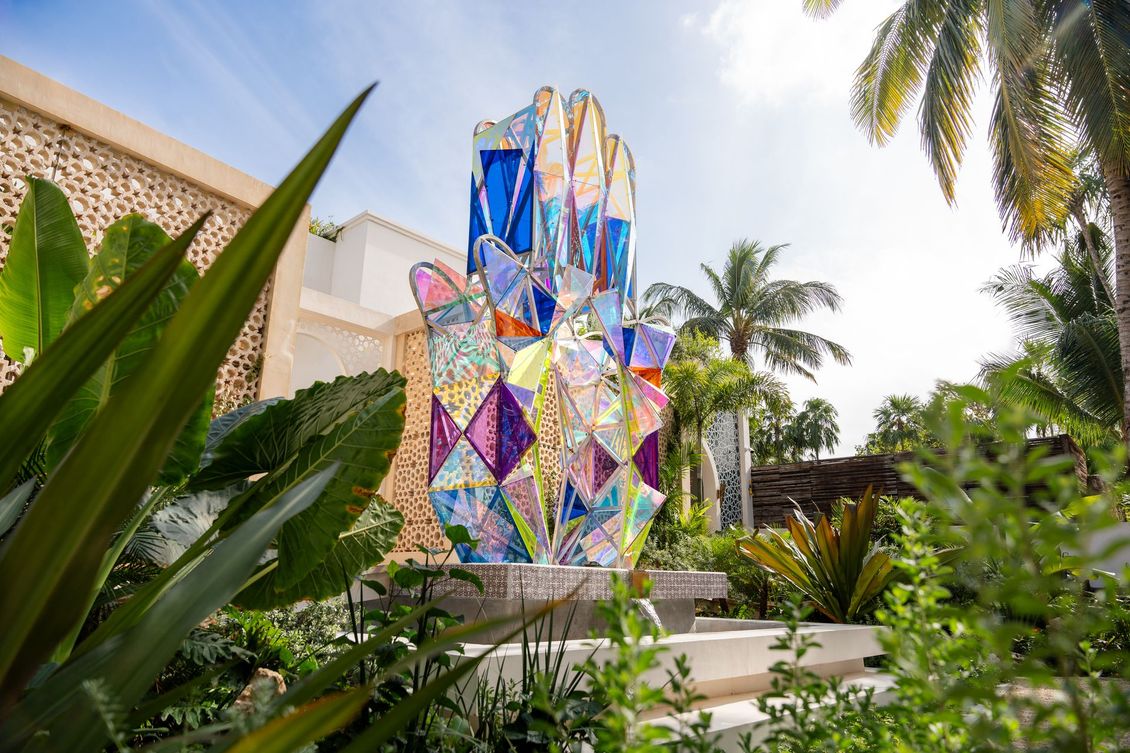 Colorful stained-glass hand sculpture in a garden with palm trees and a bright sky.
