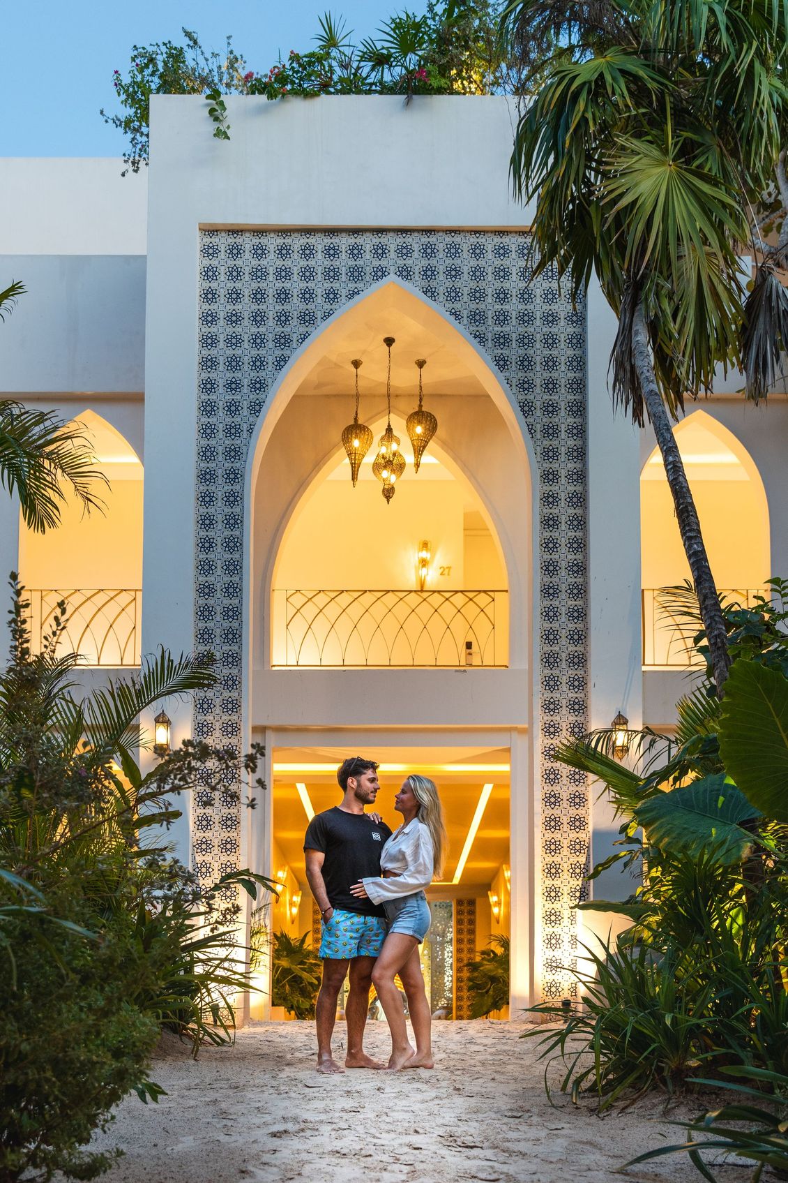 Couple stands in front of a white arched doorway, surrounded by greenery.