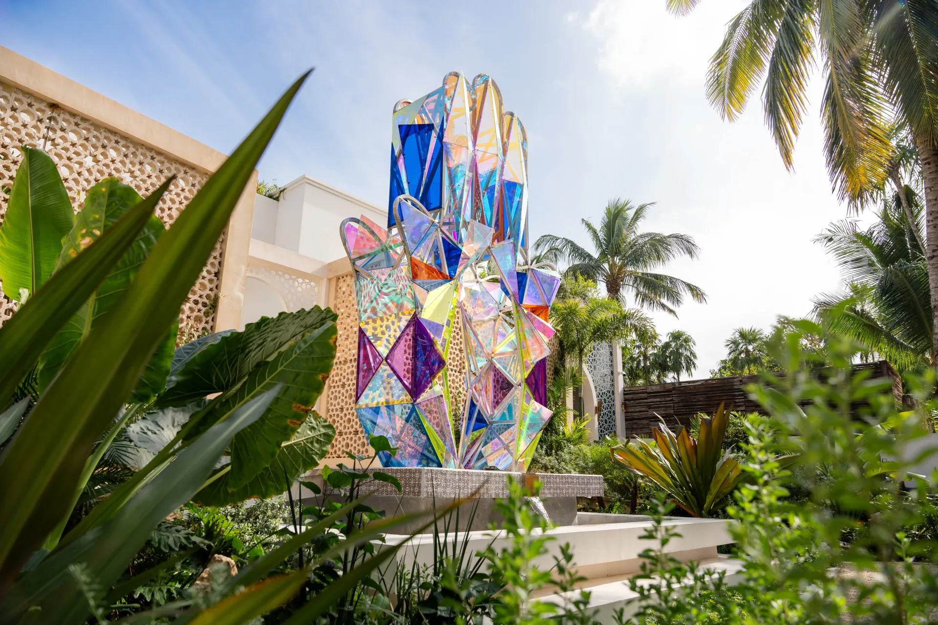 Colorful stained-glass hand sculpture in a garden, bright sunlight, surrounded by lush green plants.