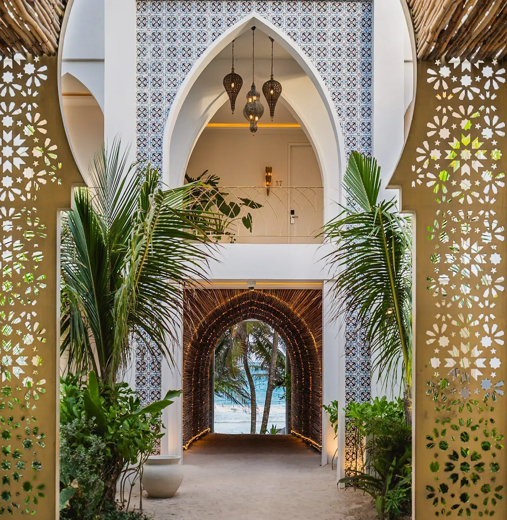 Ornate arched doorway leading to a beach. Gold latticework, lush plants, and decorative lamps.