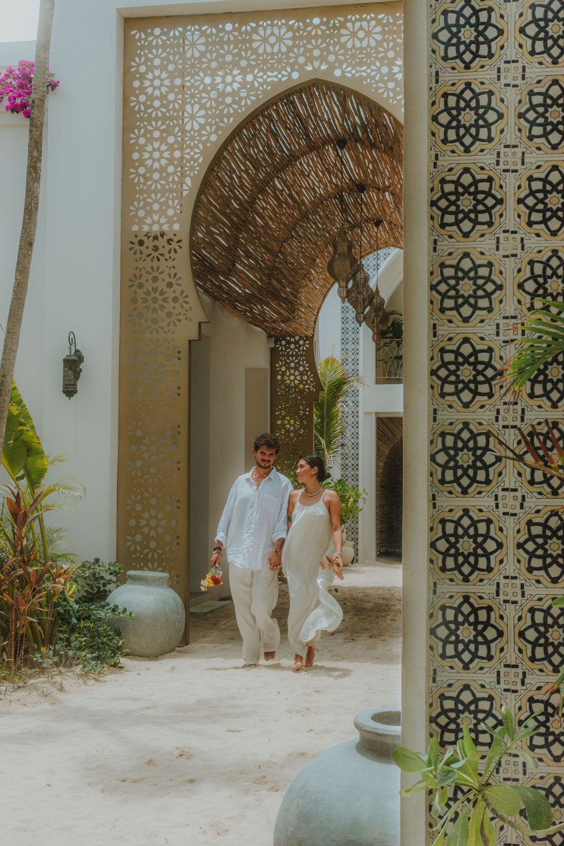 Couple walking through ornate archway, holding hands, in a courtyard with plants and decorative tile.