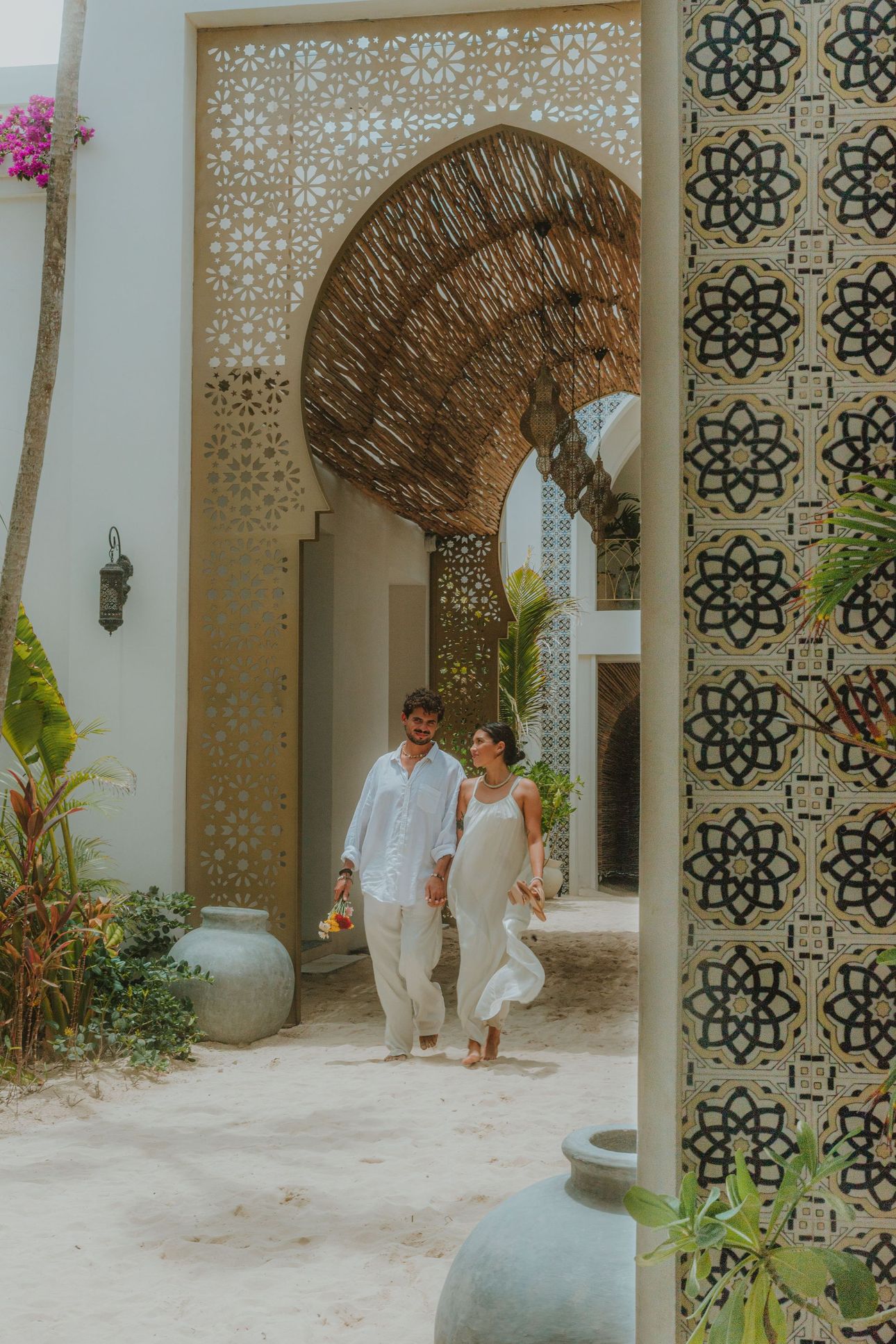Couple walking through ornate archway, holding hands, in a courtyard with plants and decorative tile.