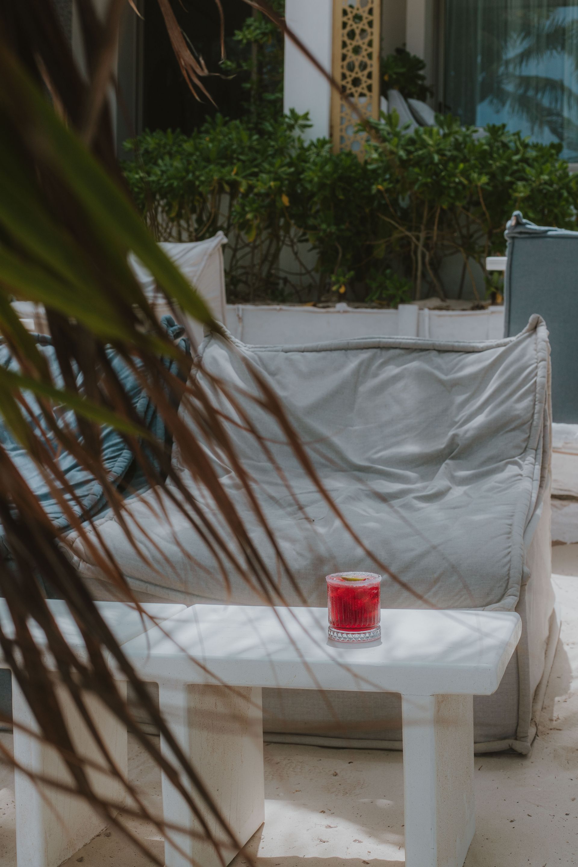 Una bebida roja reposa sobre una mesa blanca frente a un sofá blanco con almohadas azules, parcialmente oculto por una hoja de palma.
