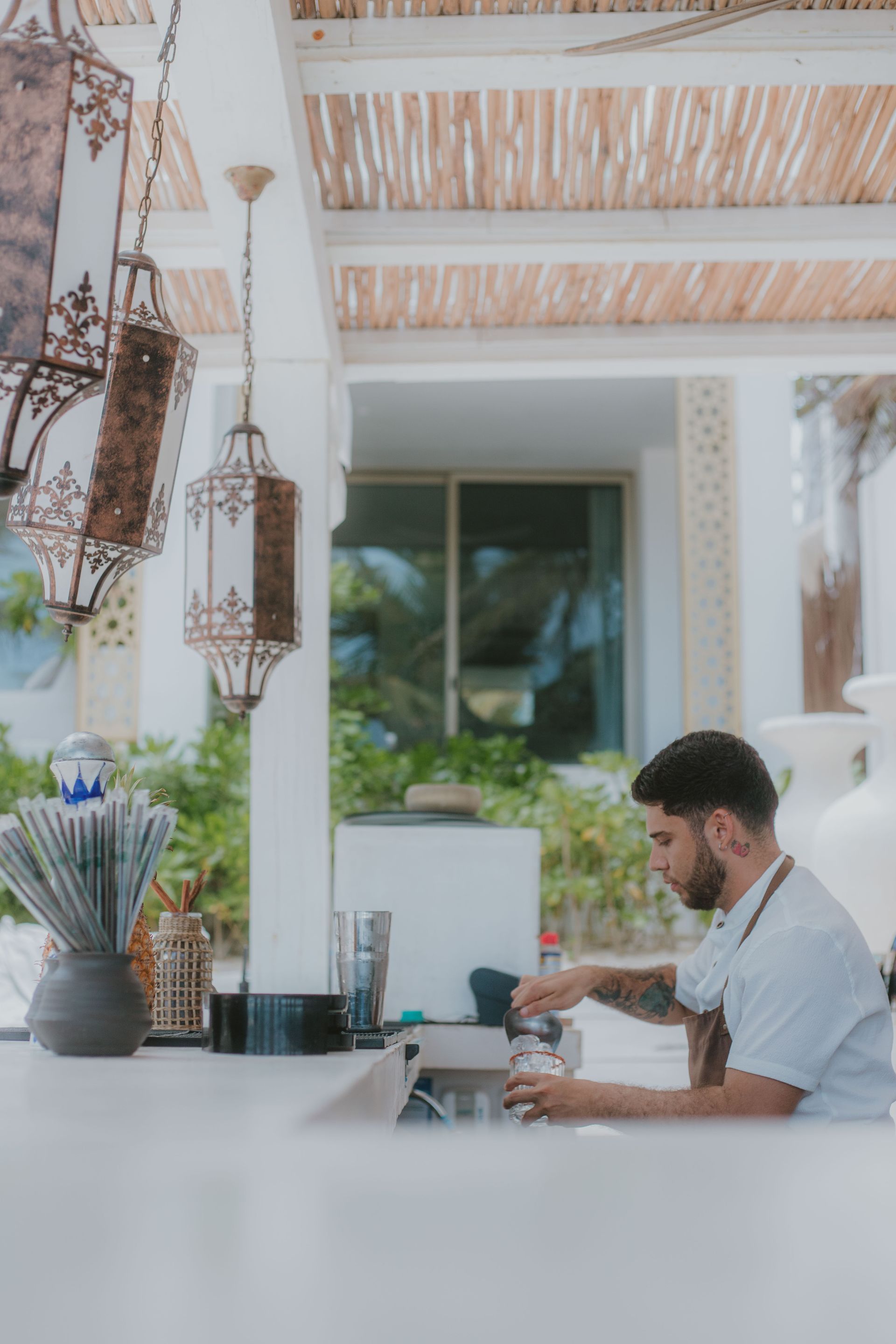 Camarero preparando una bebida en una barra blanca en un entorno al aire libre; linternas cuelgan del techo.