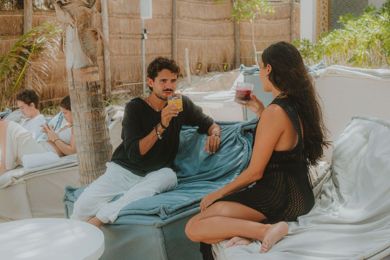 Couple toasting drinks while sitting on a cushioned bench outdoors.