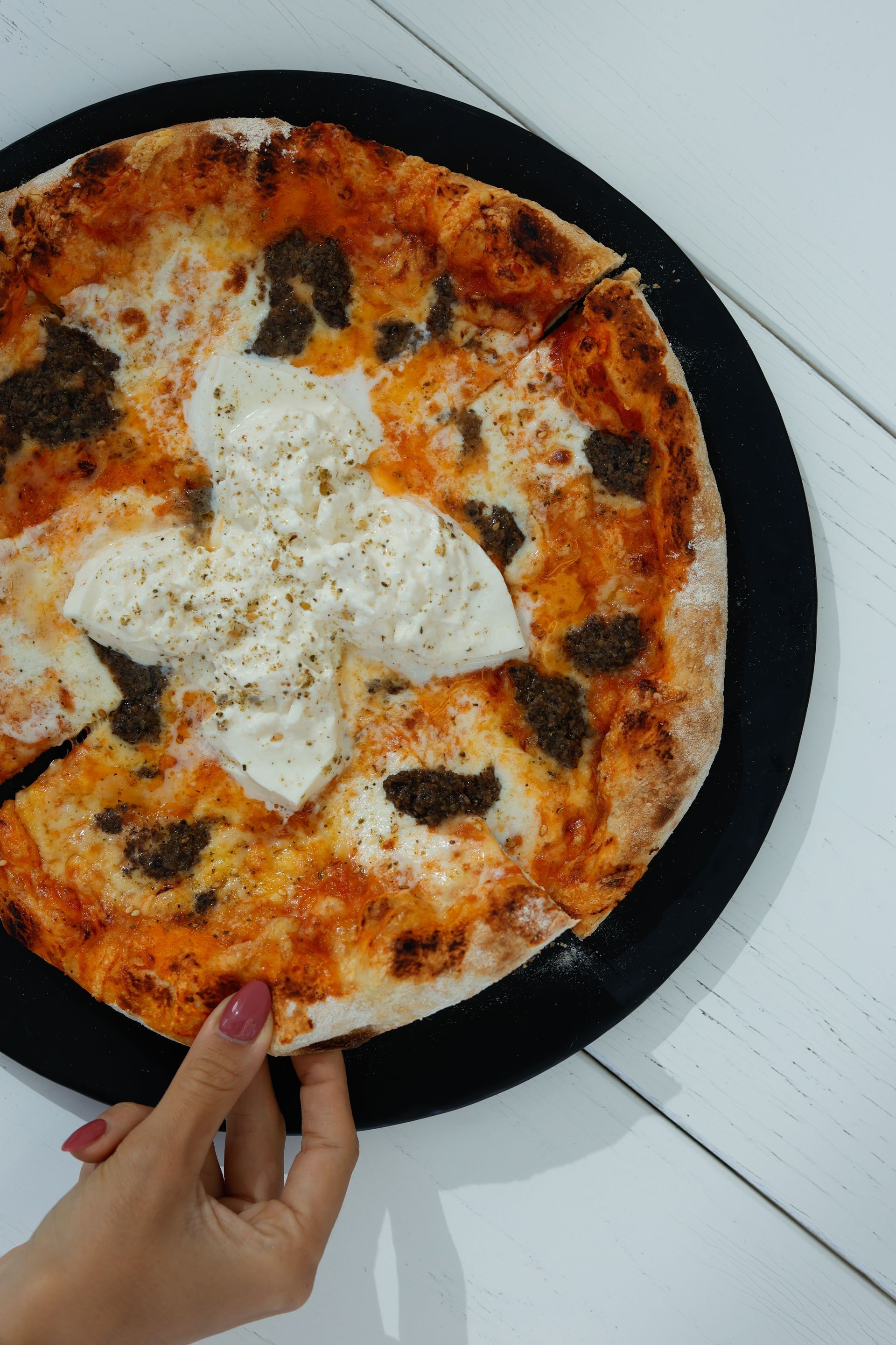 Pizza on a black plate, hand reaching to grab a slice. White cheese, pesto, and a browned crust.