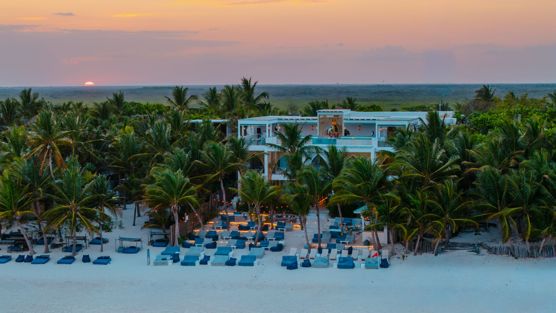 A beach resort with a white building nestled among palm trees at sunset, featuring lounge chairs on the sand.