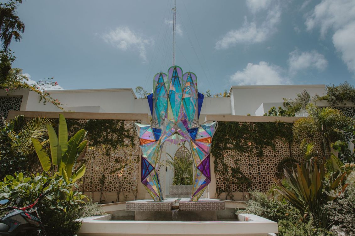 Colorful kite shaped like a hand in front of white building. Plants and a fountain surround it against a blue sky.