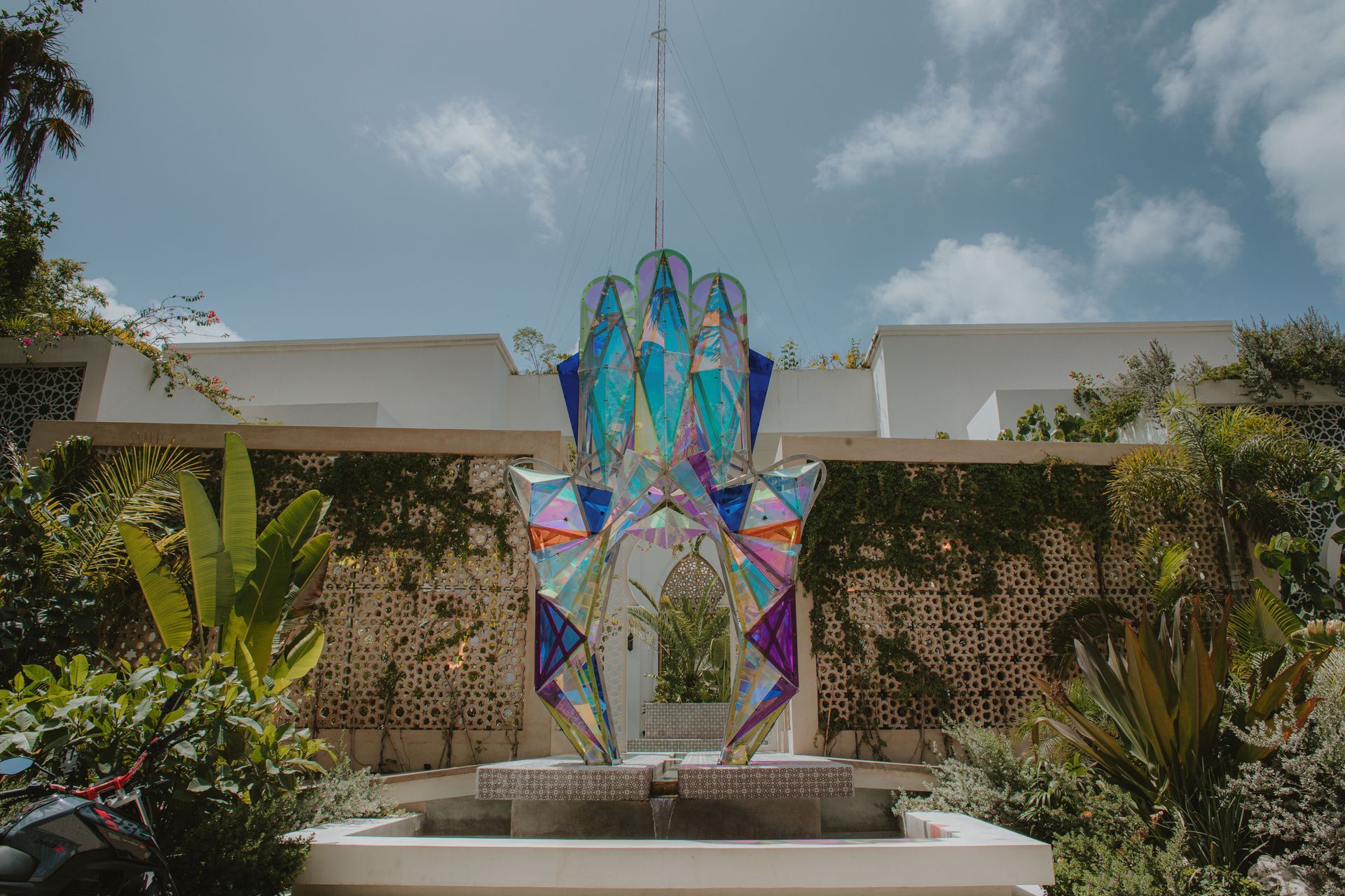 Colorful kite shaped like a hand in front of white building. Plants and a fountain surround it against a blue sky.