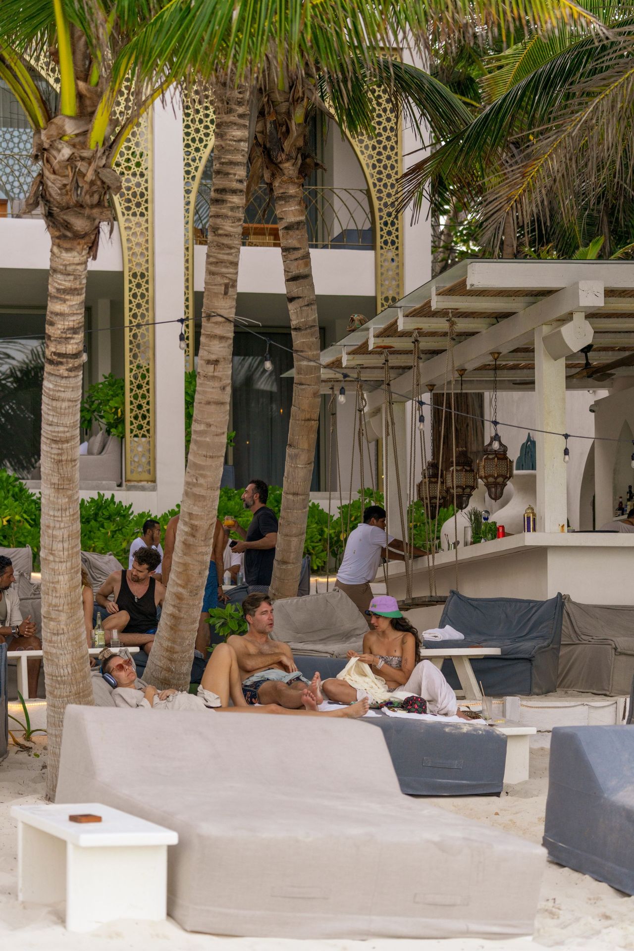 Beachside lounge area with people relaxing near a bar, white building in the background, palm trees.