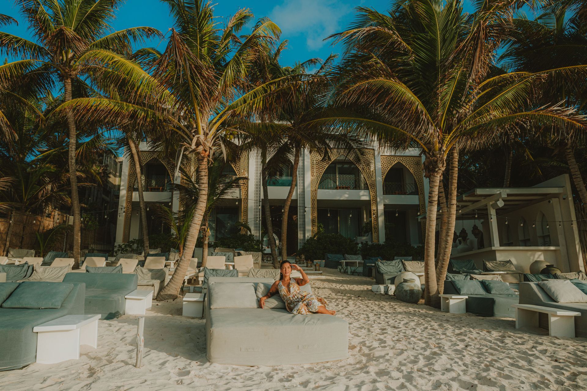 Person lounging on a gray beach bed under palm trees, facing a light-colored building.