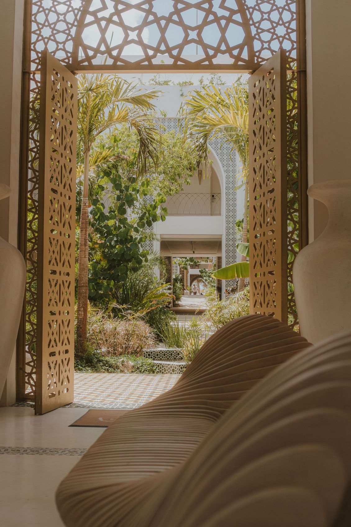 Ornate wooden doorway leading to lush garden. Sunlight filters through the intricate latticework, illuminating area.