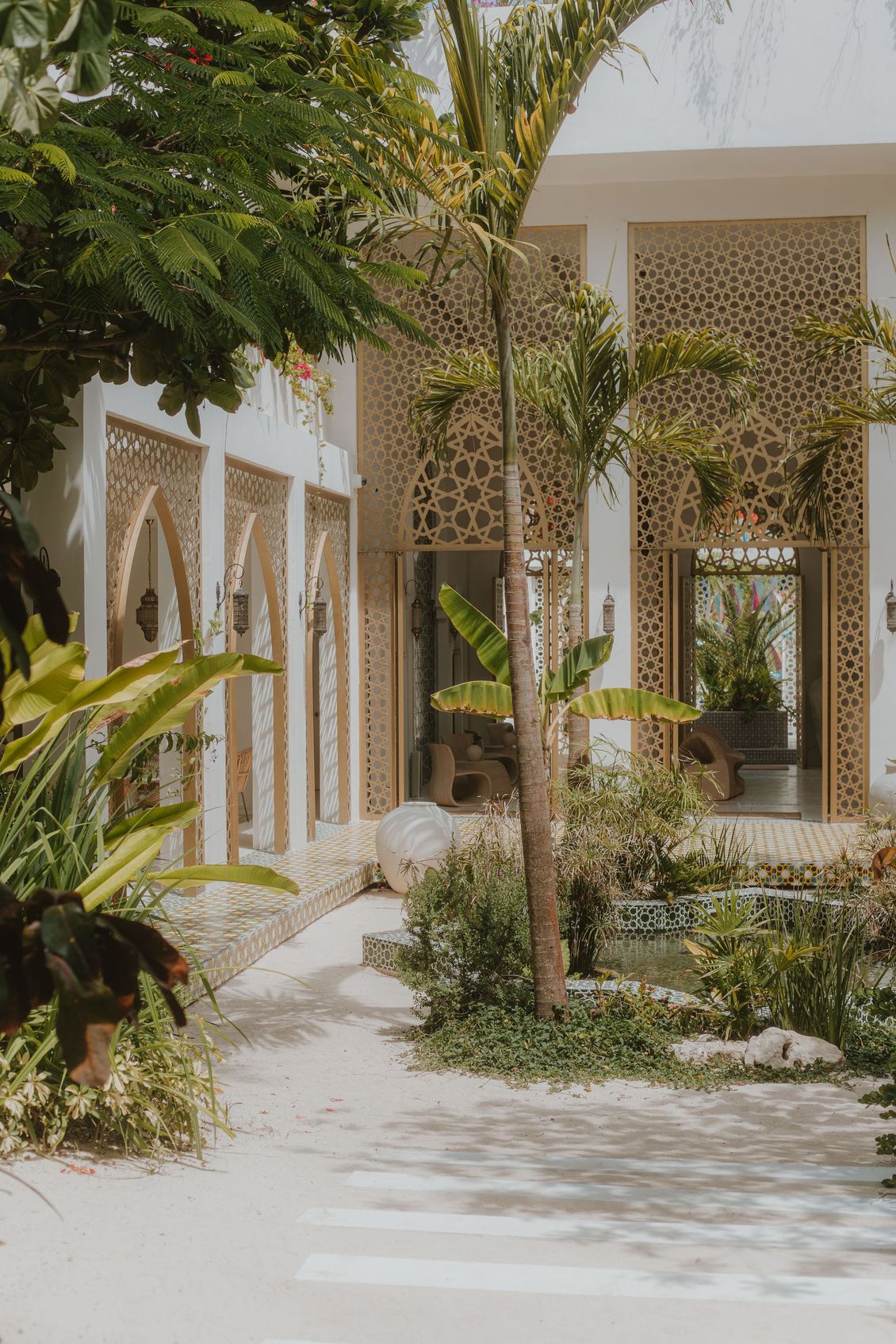 Exterior of a building with ornate gold lattice work and arched doorways, surrounded by lush greenery and a sandy path.