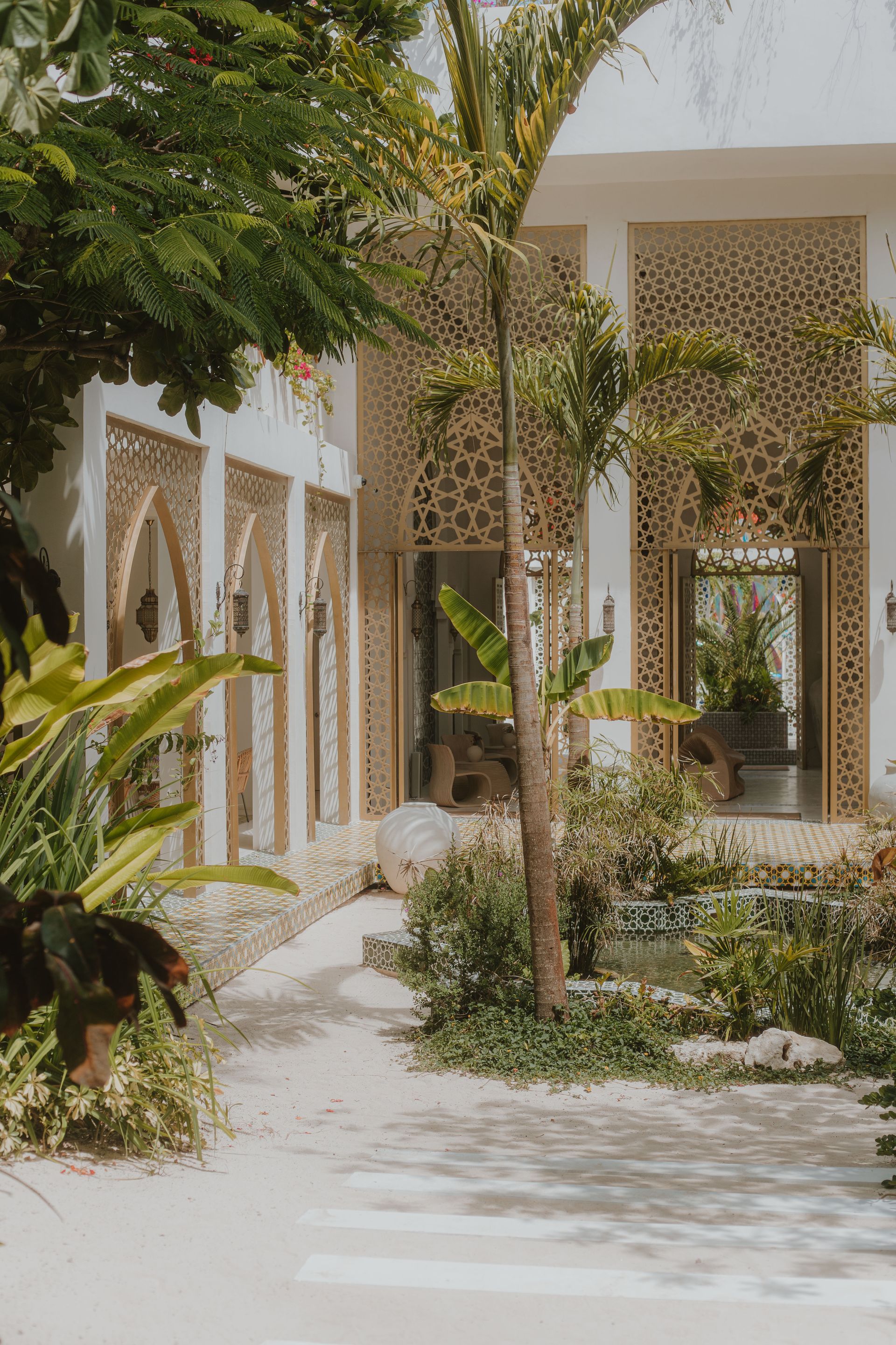 Exterior of a building with ornate gold lattice work and arched doorways, surrounded by lush greenery and a sandy path.