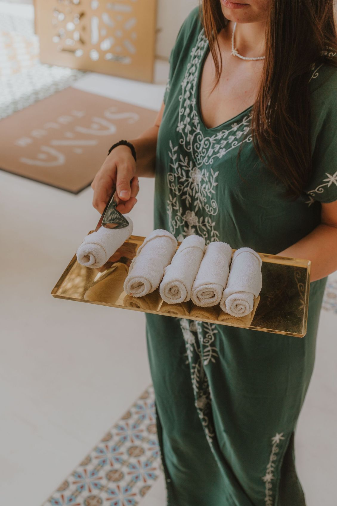 Woman holding a gold tray with five rolled white towels, wearing a green embroidered dress.